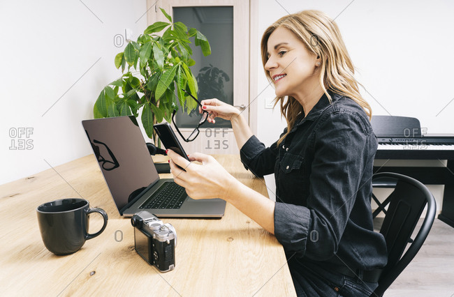 Beautiful blonde Caucasian woman works from her living room with her laptop on a wooden desk. She wears black casual clothes.