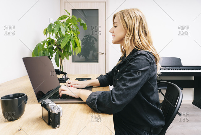 Beautiful blonde Caucasian woman works from her living room with her laptop on a wooden desk. She wears black casual clothes.