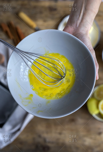 From above top view of crop anonymous woman whipping eggs in black bowl on wooden table with lemon , flour, butter and cinnamon sticks ingredients for cake