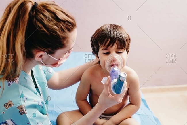 Side view of young female pediatric doctor in medical mask doing inhalation treatment with nebulizer for little boy having respiratory problem in hospital