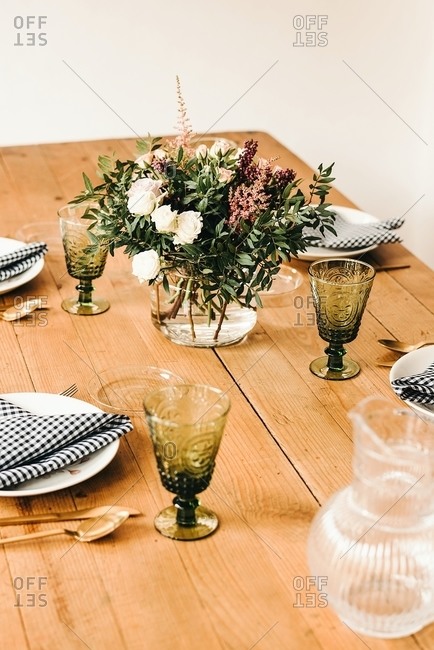 Bouquet of miscellaneous flowers and green plant twigs in vase with water on a wooden table set for a meal