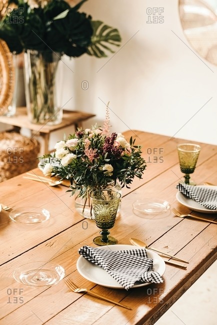 Bouquet of miscellaneous flowers and green plant twigs in vase with water on a wooden table set for a meal