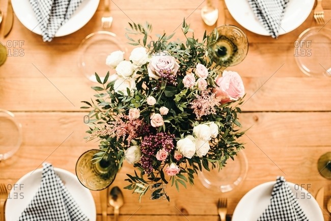 From above top view bouquet of miscellaneous flowers and green plant twigs in vase with water on a wooden table set for a meal