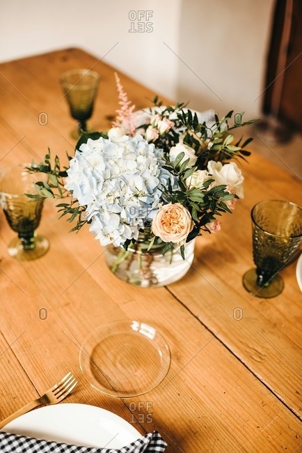 From above bouquet of miscellaneous flowers and green plant twigs in vase with water on a wooden table set for a meal