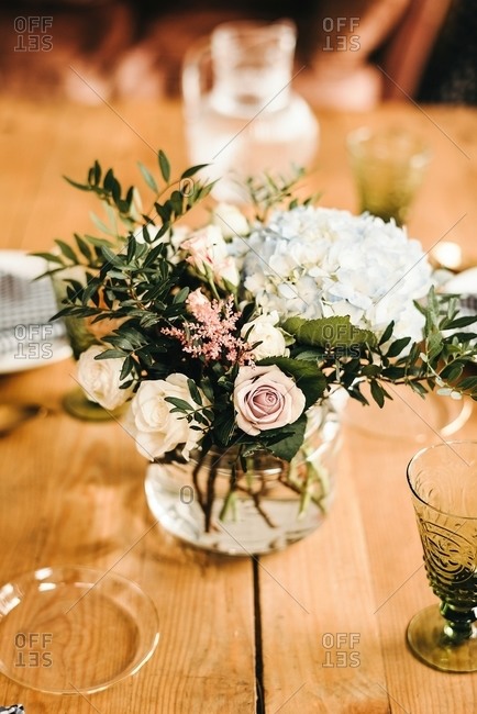 Bouquet of miscellaneous flowers and green plant twigs in vase with water on a wooden table set for a meal