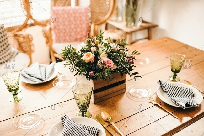 From above bouquet of miscellaneous flowers and green plant twigs in a wooden box on a timber table set for a meal with beautiful designed rattan chair on the background