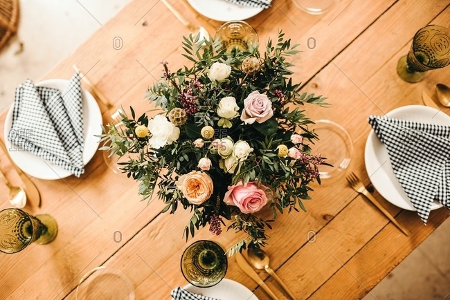From above top view bouquet of miscellaneous flowers and green plant twigs in vase with water on a wooden table set for a meal