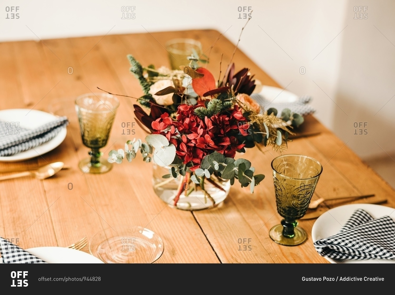 From above bouquet of miscellaneous flowers and green plant twigs in vase with water on a wooden table set for a meal