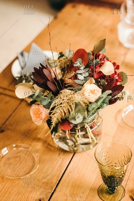 From above bouquet of miscellaneous flowers and green plant twigs in vase with water on a wooden table set for a meal