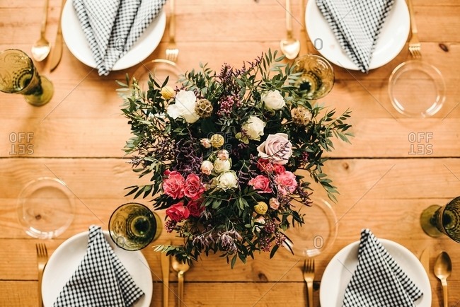 From above top view bouquet of miscellaneous flowers and green plant twigs in vase with water on a wooden table set for a meal