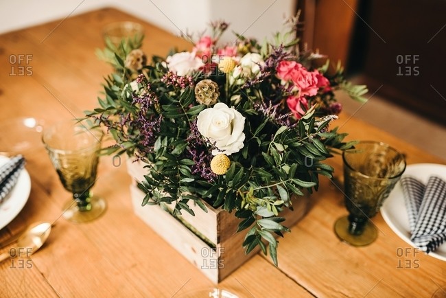 From above bouquet of miscellaneous flowers and green plant twigs in a wooden box on a timber table set for a meal