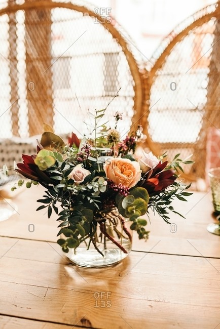 Bouquet of miscellaneous flowers and green plant twigs in vase with water on a wooden table set for a meal with beautiful designed rattan chair on the background