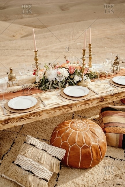 From above of long wooden table decorated with flowers and candles and served with white plates placed on carpet near traditional Arabic seats with sandy dunes in background