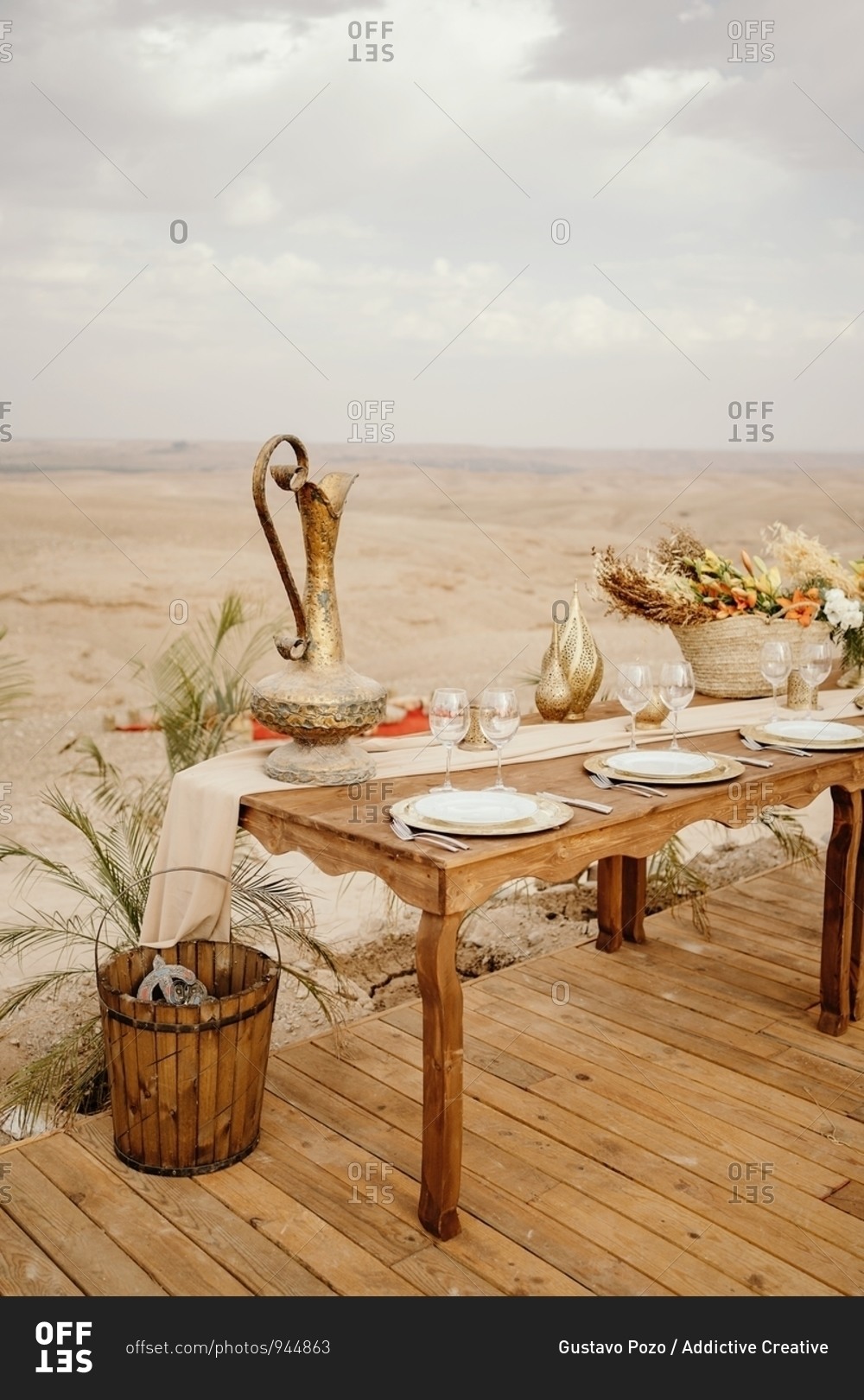 Long wooden table decorated with flowers and candles and served with white plates on traditional Arabic set with sandy dunes in background