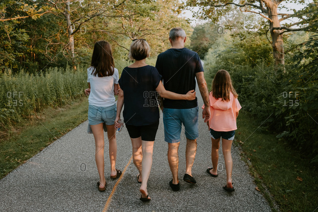 Back view of adult man and woman with daughters dressed in casual t shirts and shorts walking on pathway among green trees while spending summer day together in countryside