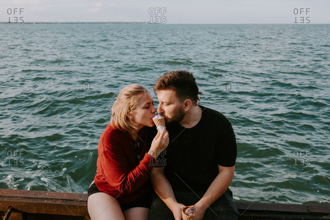Happy young romantic couple in casual wear eating ice cream cone together while sitting on pier near sea and enjoying summer holidays together