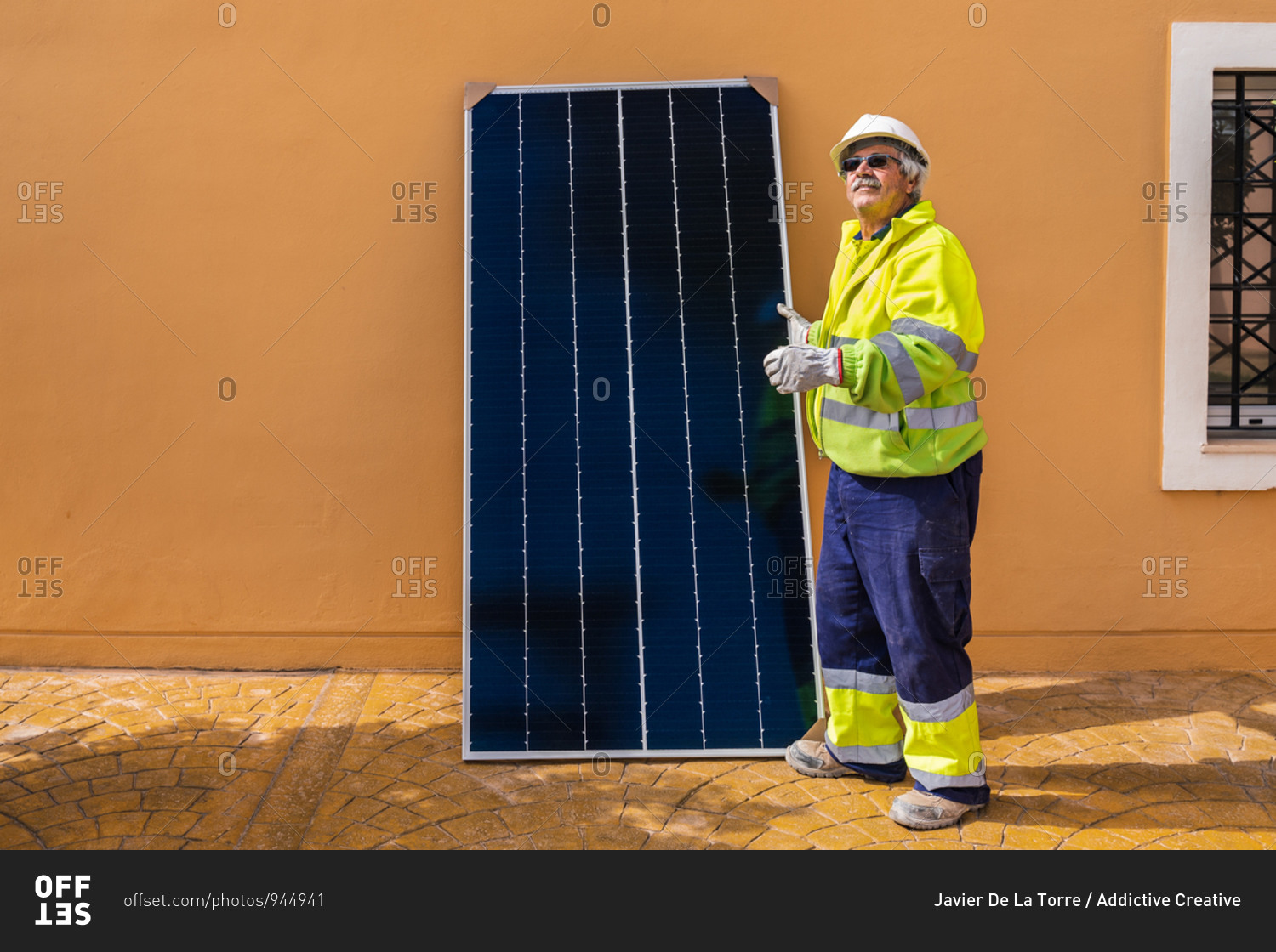 Side view of mature experienced male technician in uniform and helmet standing with solar panel near yellow building while working on installation of renewable energy system