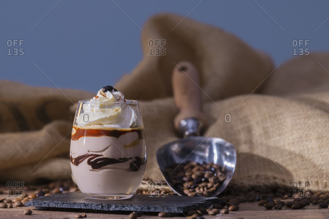 Cup with tasty parfait placed near scoop with roasted coffee beans against rough linen cloth on blue background