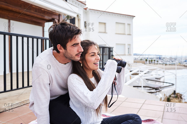 Side view of cheerful romantic couple in casual wear sitting on terrace with binoculars and enjoying view while spending holidays together at seaside