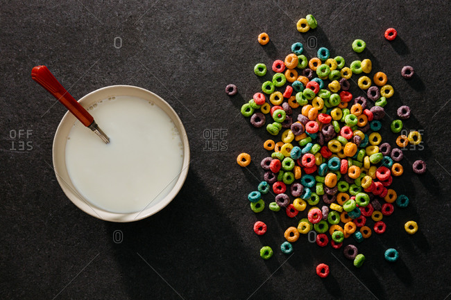 Bowl of milk with spoon next to pile of colorful cereal on table