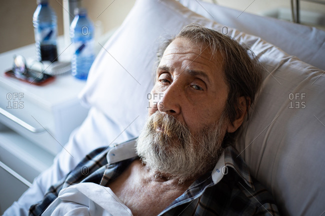 Calm aged man with beard lying under blanket on bed in hospital ward looking at camera