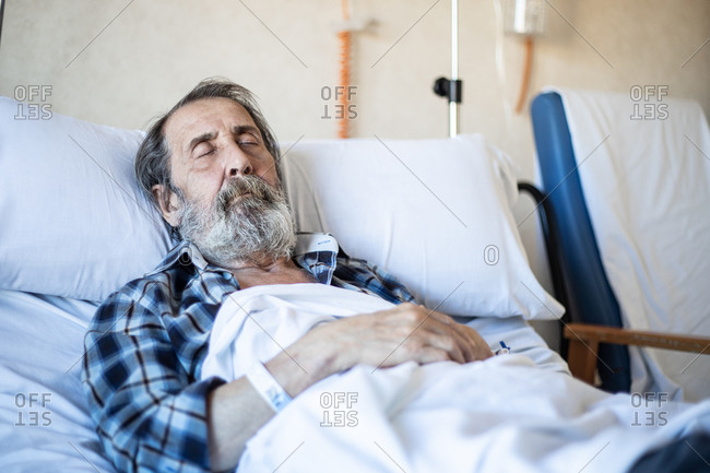 Calm aged man with beard lying under blanket on bed in hospital ward and sleeping