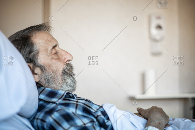 Calm aged man with beard lying under blanket on bed in hospital ward and sleeping