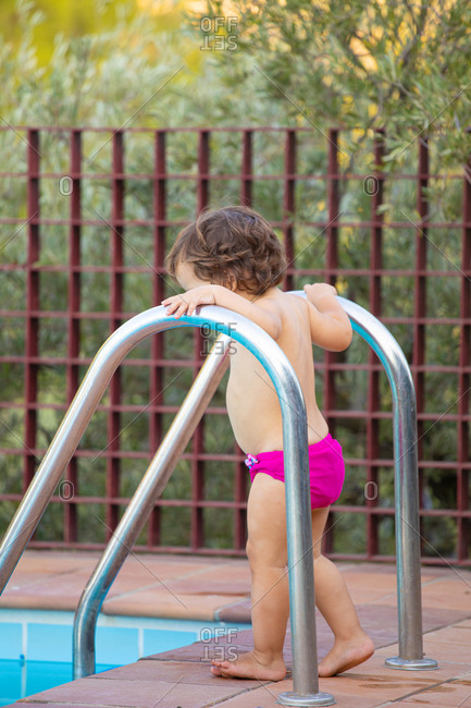 Side view of adorable shirtless toddler on poolside near metal ladder and clean blue water of pool