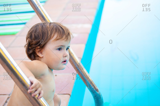 Adorable shirtless toddler looking away while sitting on poolside near metal ladder and clean blue water of pool