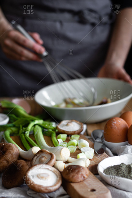 Bunch of fresh scallions and mushrooms placed on cutting board near eggs and poppy seeds against crop housewife mixing ingredients in bowl