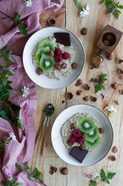 Top view of slices of fresh kiwi and raspberries placed near chocolate and hazelnuts in bowl of healthy porridge on wooden table near lilac cloth
