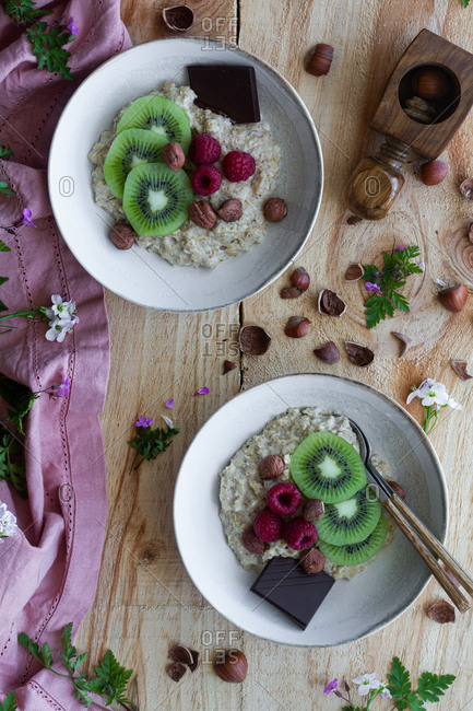 Top view of slices of fresh kiwi and raspberries placed near chocolate and hazelnuts in bowl of healthy porridge on wooden table near lilac cloth