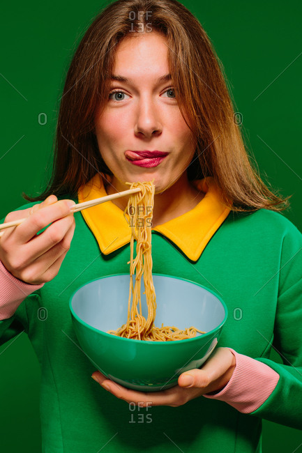 Positive young female in green shirt looking at camera grimacing sticking tongue out while eating yummy instant noodles with chopsticks on green background