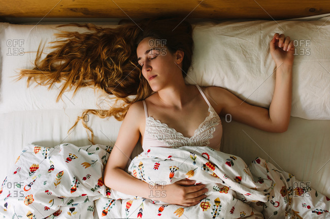 From above of young serene female with long wavy hair wearing lace bra sleeping in cozy bed with white sheets and ornamental blanket
