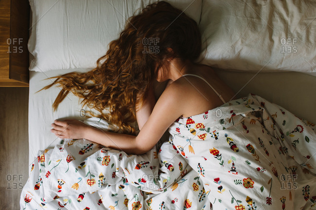 From above side view of unrecognizable serene female with long wavy hair over the face wearing lace bra sleeping in cozy bed with white sheets and ornamental blanket