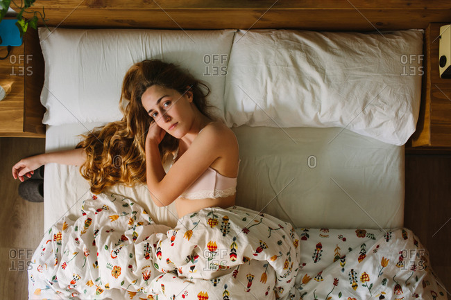 From above of young serene female with long wavy hair wearing lace bra sleeping in cozy bed with white sheets and ornamental blanket