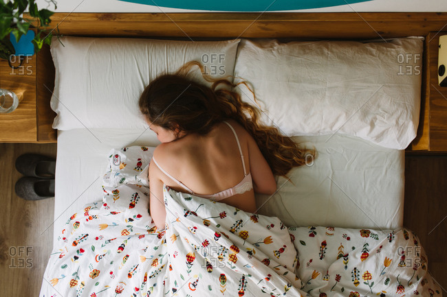 From above of young serene female with long wavy hair wearing lace bra sleeping in cozy bed with white sheets and ornamental blanket