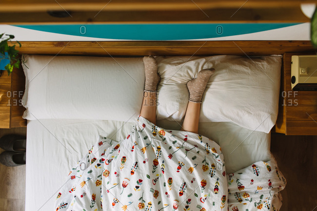 From above of crop anonymous female wearing socks lying in bed covered with blanket with legs on pillows