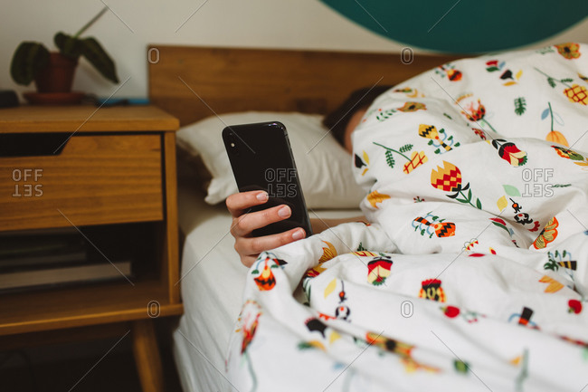 Unrecognizable female checking time on smartphone while lying under blanket in cozy bed in morning