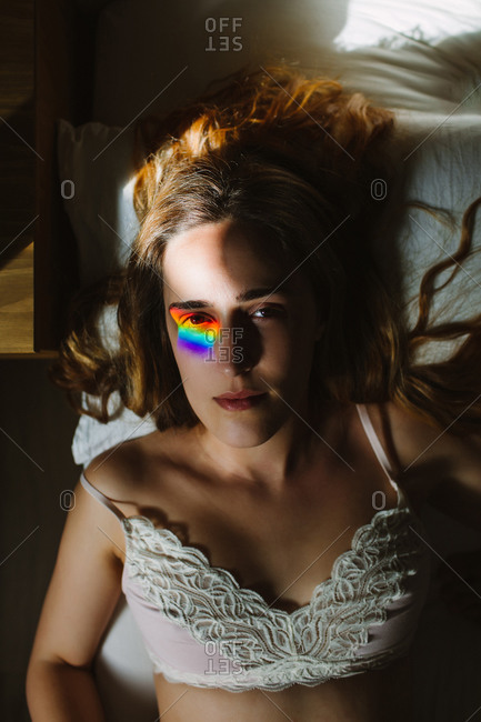From above of young sensual female in white lace bra with colorful rainbow blink on eye looking at camera while resting in bed in morning
