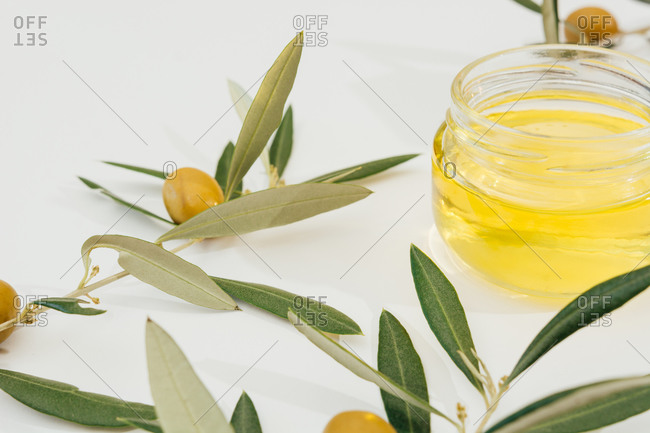 From above of natural olive oil in glass jar placed on white table with green olive tree branches