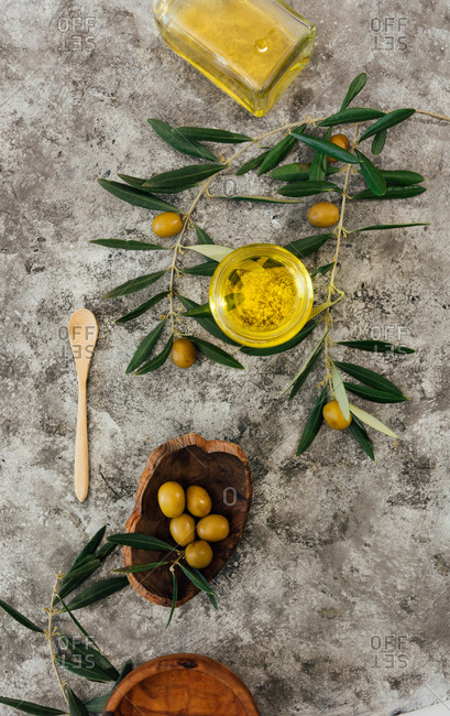 From above of glass jar with organic olive oil with wooden spoon placed on shabby gray table near fresh green olives and tree branches