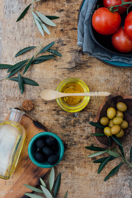 From above of glass jar with organic olive oil with wooden spoon placed on shabby gray table with fresh tomatoes green and black olives near tree branches