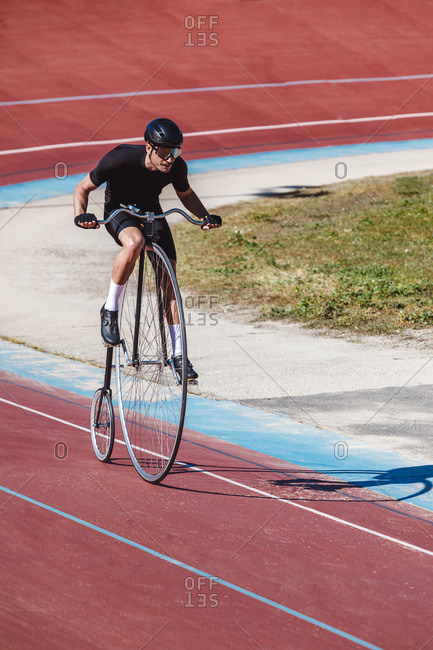 From above adult focused cyclist in black sportswear and helmet riding penny farthing along racetrack at sports stadium while getting ready for competition in sunny day