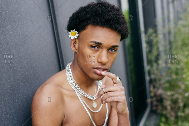 Side view alluring young black teenage man with naked torso and neck chains holding flower in mouth and looking at camera while standing against gray wall