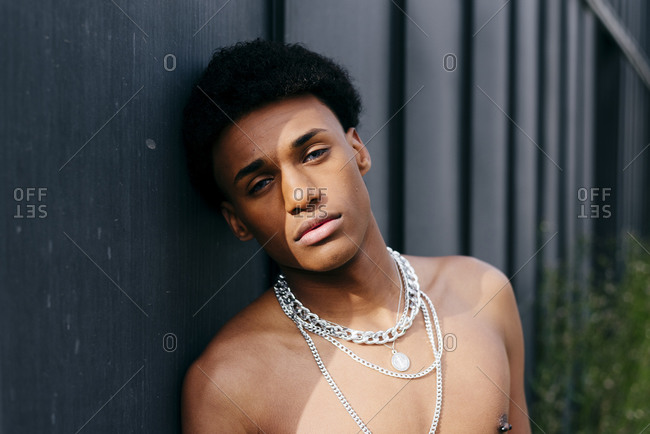Alluring young black teenage man with naked torso and neck chains holding flower in mouth and looking at camera while standing against gray wall
