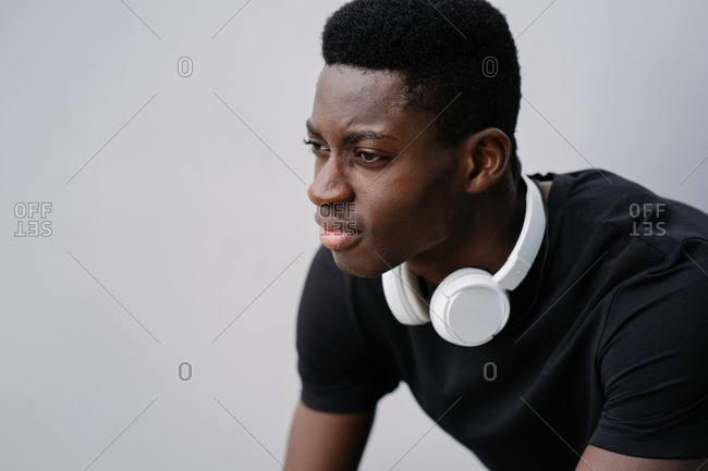 Dissatisfied African American guy with white headphones frowning and looking away while sitting against gray background on city street