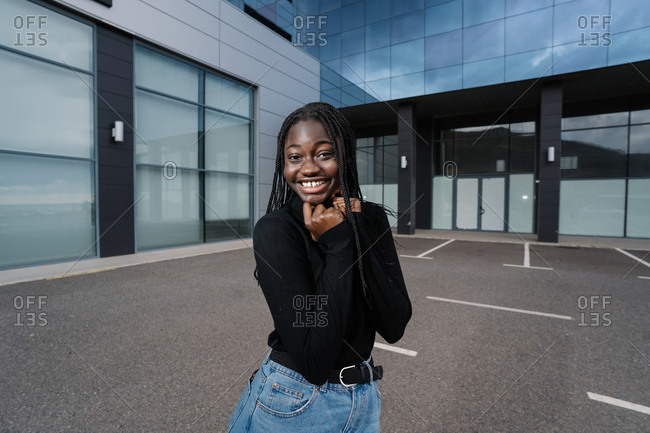 Happy black female in casual clothes cheerfully smiling and looking at camera while standing on parking lot outside modern building on city street