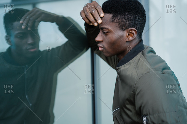 Side view of black guy in casual outfit touching face and looking at reflection on glass wall while spending time on modern city street
