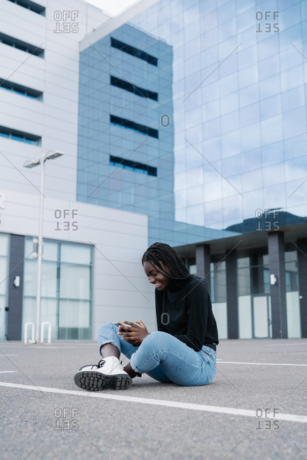 Full body delighted black female in casual clothes smiling and browsing smartphone while sitting on parking lot with crossed legs outside modern building on city street